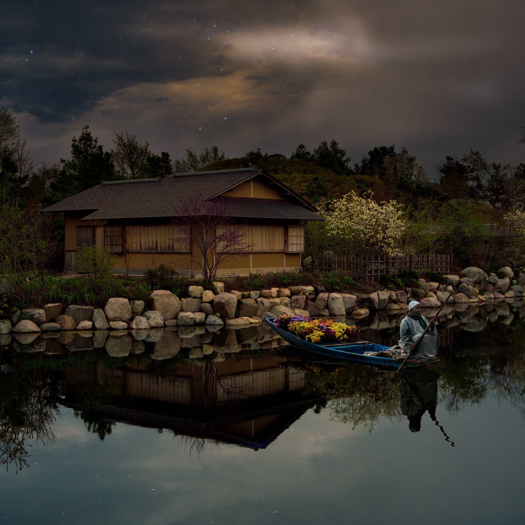 man sitting in boat on still water with small home on shore behind him